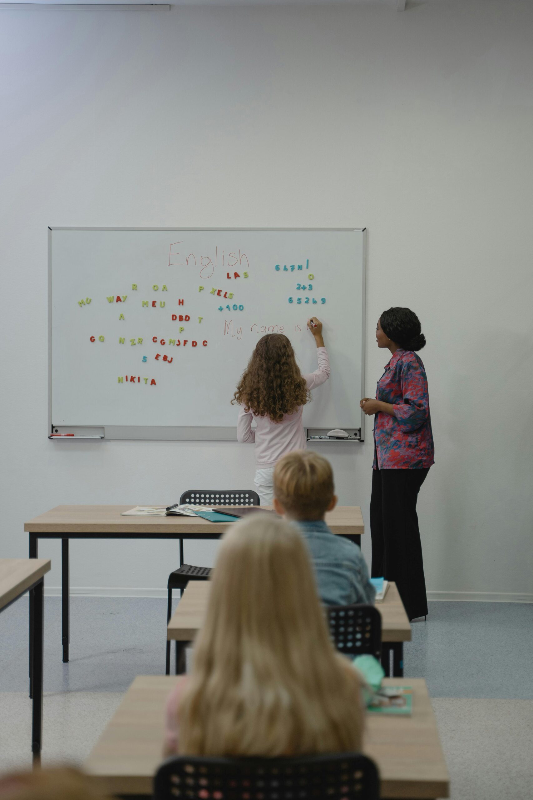 Teacher instructing diverse students with magnetic letters on a classroom whiteboard.
